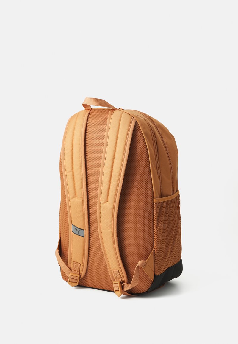 Mochila de tela naranja con respaldo de malla acolchada, correas ajustables, un parche con logotipo en la parte frontal y base negra. Cuenta con bolsillos laterales para almacenamiento.