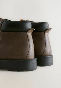 Et par brune læderstøvler med sorte polstrede kraver og tykke sorte gummisulder, set fra bagsiden mod en lys baggrund.