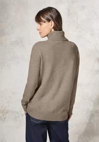 Beige, oversize stickad tröja med hög polokrage och ribbad fåll. Modell står med händerna i fickorna och visar upp den avslappnade passformen.