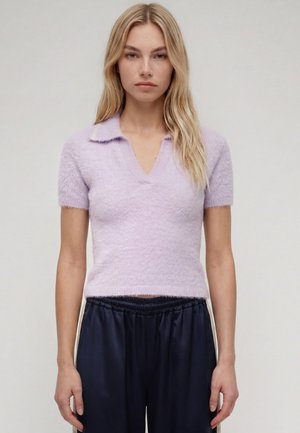 Femme blonde portant un haut violet clair duveteux à manches courtes avec col en V et un pantalon noir ample, debout devant un fond uni.