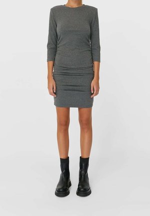 Femme portant une robe grise ajustée au genou avec des manches trois-quarts et des bottines noires, debout devant un fond uni.