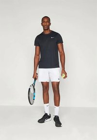 Nike svart kortärmad sporttröja, vita träningsshorts, svarta sneakers, håller en tennisracket och boll framför en enkel bakgrund.