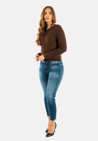Bruin textuurtrui met ronde hals, knoopsluiting, gecombineerd met blauwe slim-fit jeans en zwarte hoge hakken.