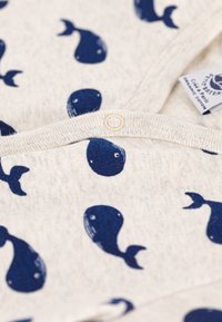 Organinis medvilninis drabužis su kreminiu pagrindu ir tamsiai mėlynais banginių raštais, pabrėžtas centrinės sagos ir minkštos tekstūros.