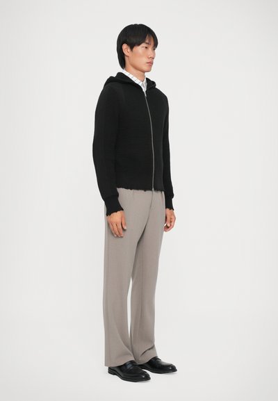 Svart stickad zip-hoodie med en strukturerad yta och huva, kombinerad med ljusgrå byxor och svarta skor, stylad mot en neutral bakgrund.