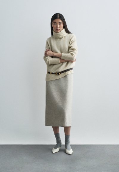 Μπεζ πλεκτή ζακέτα με ψηλό γιακά, συνδυασμένη με μπεζ φούστα μέχρι το γόνατο. Φορεμένη με γκρι κάλτσες και λευκά παπούτσια με μυτερή μύτη. Καφέ ζώνη με χρυσές λεπτομέρειες.