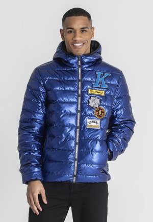 Blauwe puffy jas met een glanzende afwerking, voorzien van meerdere kleurrijke patches en een rits aan de voorkant, aangevuld met een capuchon.