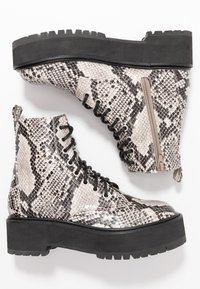 Un par de botines de plataforma con estampado de serpiente en blanco y negro, cordones negros, cremalleras laterales y suelas gruesas en un fondo blanco.