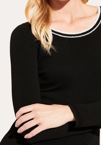 Haut noir à manches longues côtelé avec un col rond et des bordures blanches, mettant en valeur une coupe ajustée et une texture douce. Main reposant sur le bras.