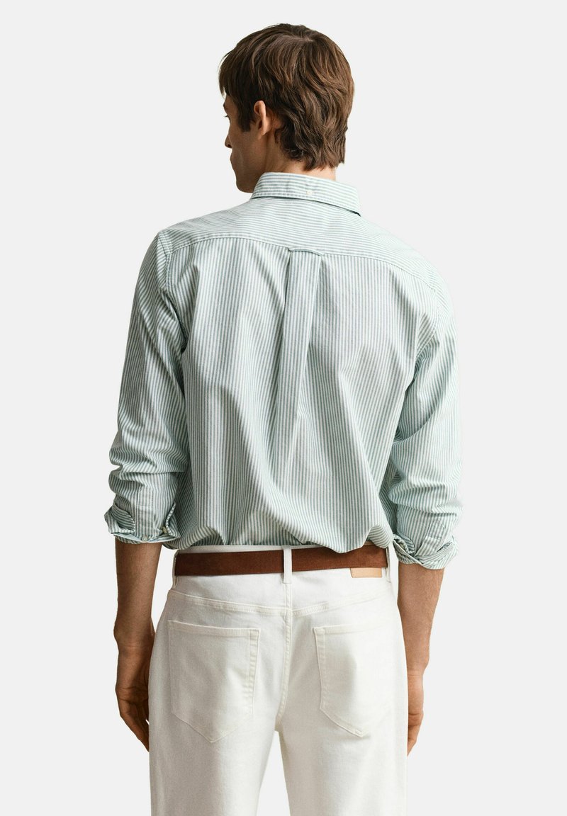 Uomo di spalle che indossa una camicia a maniche lunghe a righe bianche e verde chiaro con le maniche arrotolate, pantaloni bianchi e una cintura marrone.