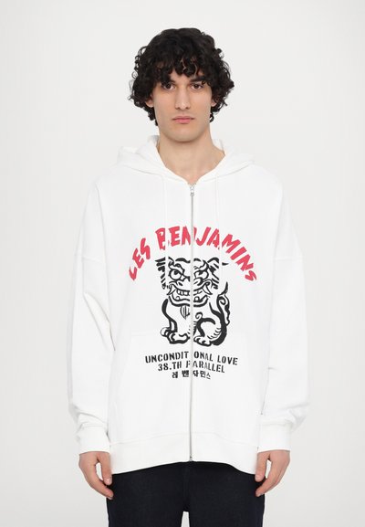 Veste à capuche blanche zippée avec un design graphique représentant un tigre noir et du texte. Poche kangourou, coupe ample, tissu décontracté et capuche ajustable.