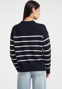 Femeie cu păr lung și închis la culoare poartă un pulover bleumarin cu dungi orizontale albe și blugi din denim deschis, întorcându-se cu fața la un fundal simplu.