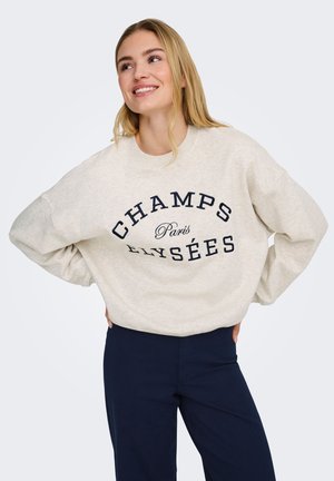 Lachende blonde vrouw die een beige sweatshirt draagt met de tekst "CHAMPS Paris ELYSÉES" en marineblauwe broek, handen in de zij, kijkt omhoog naar rechts.