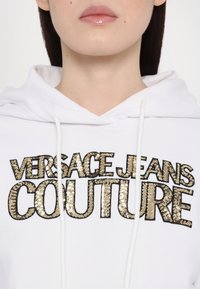 Felpa bianca con ricamo in paillettes dorate e nere che recita "VERSACE JEANS COUTURE". Il materiale sembra morbido, testurizzato, con dettagli a cordoncino.