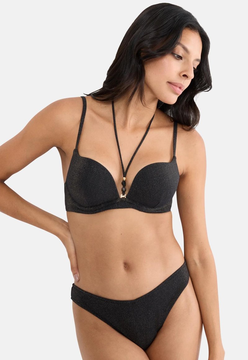 Γυναίκα που φοράει μαύρο μπικίνι με γκλίτερ και κολιέ σε στυλ λαιμού, ποζάροντας με το ένα χέρι στη μέση της, κοιτώντας προς τα κάτω.