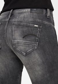 Jeans de mezclilla negros deslavados con un primer plano del bolsillo trasero, que presenta un diseño curvado cosido y un parche de cuero en la cintura.