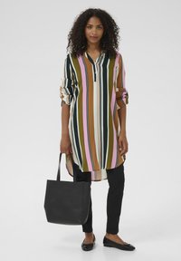 Tunique rayée avec des lignes verticales vertes, roses, brunes et noires, patte de boutonnage à l'avant, accompagnée de pantalons noirs et d'un sac fourre-tout noir.