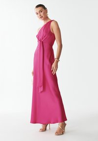 Fuksijos rožinė, be rankovių suknelė su susuktos mazgo detale ties liemeniu ir plazdančia siluetu, derinama su auksinėmis šlepetėmis.