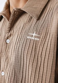 Camisa marrón con textura y pliegues verticales, con cuello puntiagudo, cierre de botones blancos y un pequeño logo bordado en blanco.