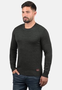 Pull en maille vert foncé avec un col rond et des manches longues. Il présente des poignets et un ourlet côtelés, avec un petit logo brodé en bas à gauche.