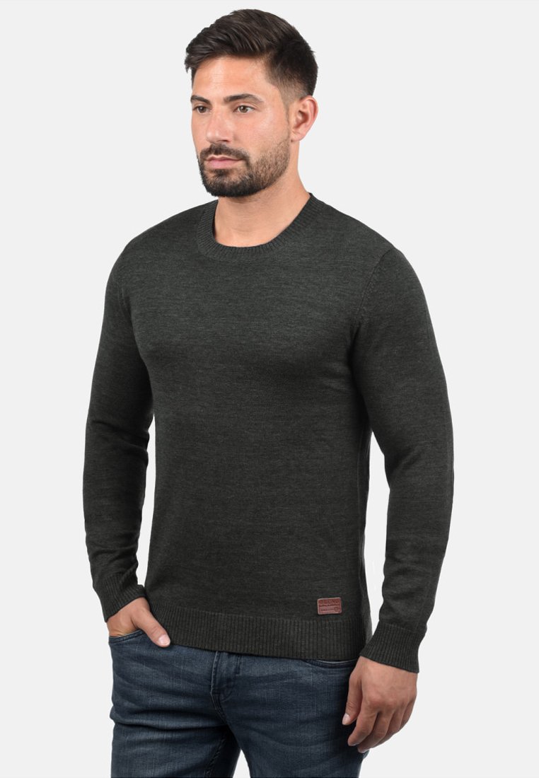 Pull en maille vert foncé avec un col rond et des manches longues. Il présente des poignets et un ourlet côtelés, avec un petit logo brodé en bas à gauche.