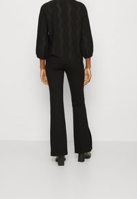 Blusa nera textured con un motivo a onde e maniche a sbuffo abbinata a pantaloni neri a zampa d'elefante, mostrata da dietro. Tacchi neri piatti visibili.