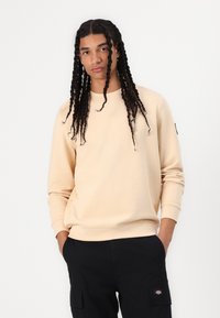 Beige sweatshirt with a crew neck and long sleeves, featuring ribbed cuffs and a logo patch on the left sleeve. Textured fabric.
