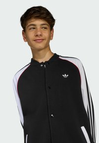 Crno-bijela jakna s kopčanjem na dugme, rebrastim ovratnikom i crvenim detaljima. Sadrži mali bijeli logo na prsima.