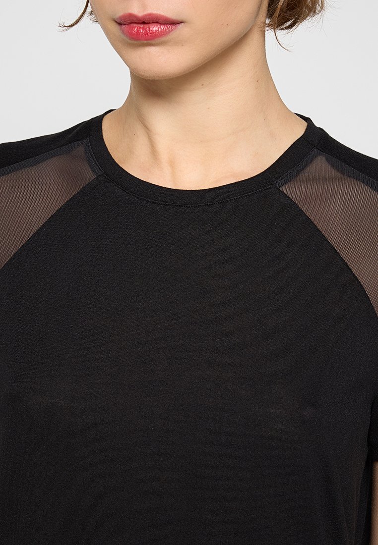 Mujer vistiendo una camiseta negra con paneles de malla transparente en los hombros, lápiz labial rojo visible y mechones de cabello corto cerca del cuello.