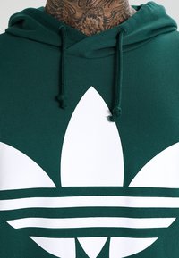 Grön huvtröja i ribbat tyg med en stor vit Adidas treflon-logo och tre horisontella ränder längst ner.