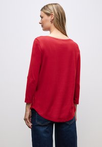 Rotes Langarmoberteil mit Rundhalsausschnitt und abgerundetem Saum, aus weichem Stoff hergestellt. Getragen mit dunkelblauen Jeans, die eine lässige Passform und seitliche Schlitze zeigen.