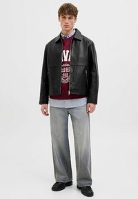 Schwarze Lederjacke mit hohem Kragen, Frontreißverschluss, zwei Seitentaschen, getragen über einem weinroten Sweatshirt, kombiniert mit hellgrauen weiten Hosen.