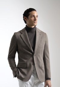 Blazer a cuadros marrones con un diseño estructurado, combinado con un suéter de cuello alto oscuro. Tejido suave, corte ajustado y dos bolsillos frontales.