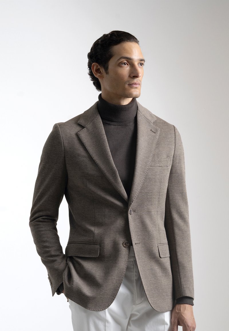 Blazer a cuadros marrones con un diseño estructurado, combinado con un suéter de cuello alto oscuro. Tejido suave, corte ajustado y dos bolsillos frontales.