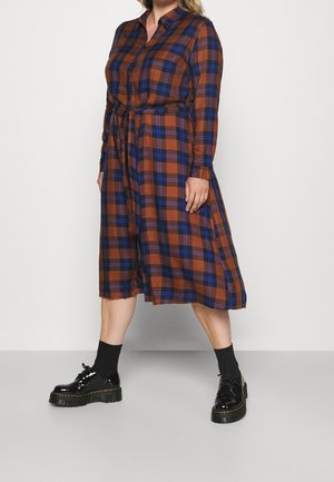 Kobieta ubrana w brązowo-niebieską kratkowaną sukienkę midi z długimi rękawami i paskiem w talii, do tego czarne skarpetki i błyszczące czarne buty na platformie.