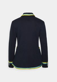 Námořnický blazer se přiléhavým střihem, s kontrastním modrým a žlutým lemováním kolem límce a spodního okraje, a s knoflíkovými detaily na rukávech.
