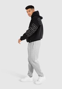 Černá mikina s kapucí a bílým nápisem "SM" na rukávech, doplněná šedými tepláky a bílými teniskami. Jednoduchý, neformální sportovní outfit.