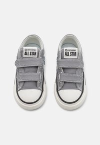 Sive platnene tenisice s bijelim gumenenim vrhom, čičak trakom i crnim bočnim detaljima. Imaju oznaku "Converse All Star" na jeziku.