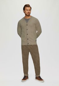 Helle graue Strickjacke mit V-Ausschnitt, sechs schwarzen Knöpfen, gerippten Bündchen, kombiniert mit braunen Cordhosen und braunen Turnschuhen. Einfarbiger Hintergrund.