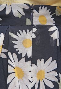 Tkanina sukienki w kwiaty ma żółte stokrotki na ciemnym tle. Faldowane wykończenie z widocznym zamkiem z tyłu. Teksturowana powierzchnia.