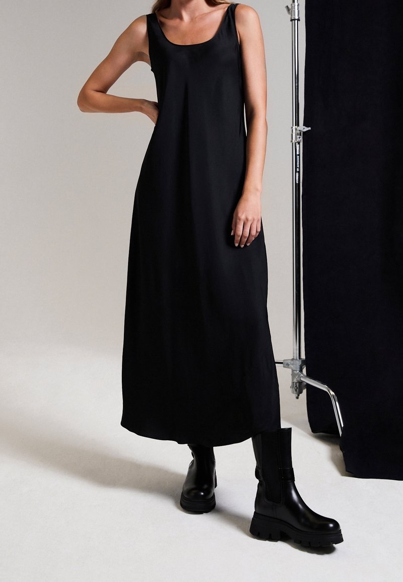 Femme portant une robe longue noire sans manches et de grosses bottes noires, debout une main sur la hanche dans un studio minimaliste.