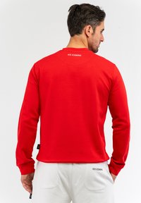 Felpa rossa a maniche lunghe con scollatura a girocollo. Presenta il logo sul retro del collo e un'etichetta laterale. Tessuto morbido e design aderente.