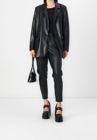 Zwarte faux leren blazer gecombineerd met zwarte faux leren broek. Hoge platformschoenen met banddetails, aangevuld met een kleine zwarte handtas.