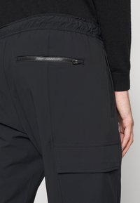 Pantalon en tissu technique noir avec une ceinture élastique, une poche arrière zippée, une poche cargo latérale et une texture lisse.