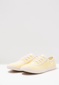 Des baskets en toile jaunes présentent un design bas, une pointe arrondie, des lacets blancs et une semelle texturée avec des accents nervurés.
