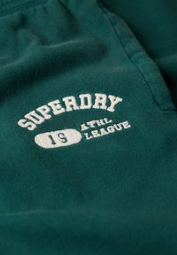 Teal stof med blød tekstur, der har hvidt broderet tekst: "SUPERDRY 19 ATHL LEAGUE" i et fedt, buet design.