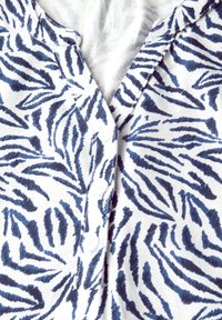 Kortærmet button-up skjorte i hvid med blåt zebrastriber mønster. Har en V-udskæring og tre små hvide knapper foran.