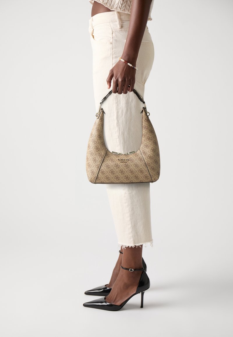 Femme tenant un sac à main Guess beige à motifs, portant un pantalon crème, un top court en maille et des talons hauts pointus noirs.