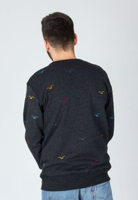 Uomo in piedi con la schiena rivolta, che indossa un maglione grigio scuro con un piccolo ricamo di un uccellino colorato e dei jeans azzurri chiari.