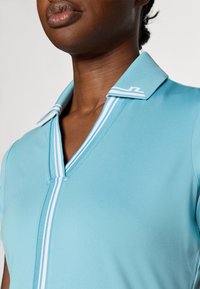 Světle modré polo tričko s výstřihem do V a kontrastními proužky na límci. Vyrobeno z hladkého, elastického materiálu. Žádné viditelné vzory ani ozdoby.